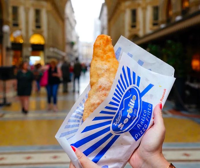 Hands lifting a pizza frita, emerging from a branded paper sleeve, set against the backdrop of an ornate shopping arcade.