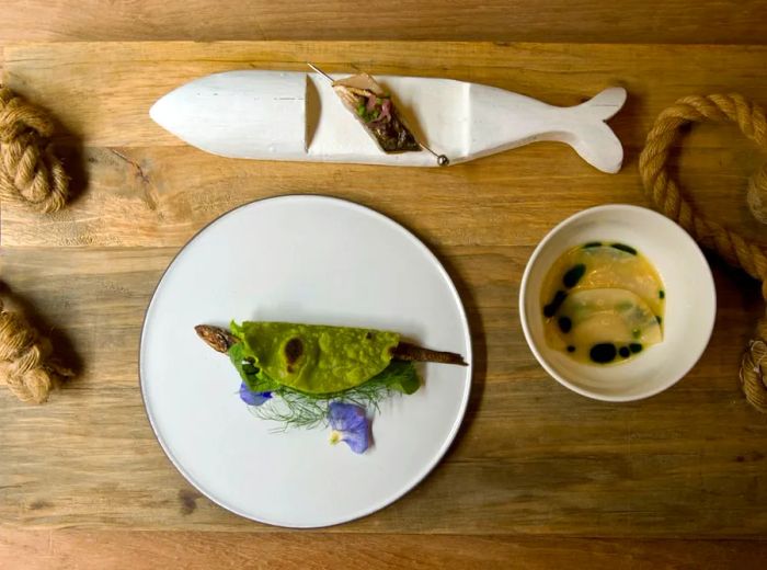 A wooden board with rope handles displays three dishes: a fish wrapped in green, skewered fish topped with pickled vegetables, and dumplings drizzled with dark sauce