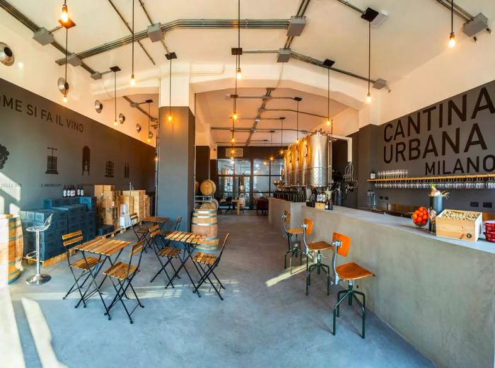 The winery's interior features sleek concrete floors and counters, with exposed bulb pendant lights illuminating patio tables and chairs, alongside large metal wine vats at the back.