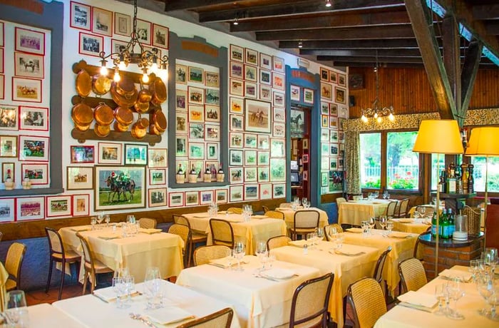 A cozy restaurant interior featuring white tablecloths, cane chairs, an array of pendant lights, and a large wall adorned with numerous small pictures and artworks.