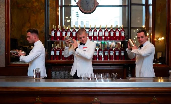 Bartenders dressed in white tuxedo jackets vigorously shake cocktail shakers in front of a back bar filled with an array of Campari bottles.