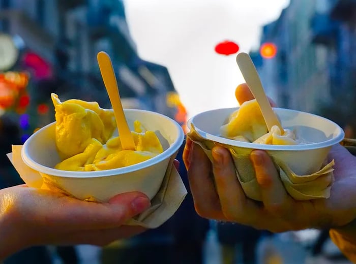 Two individuals hold paper cups filled with dumplings, each with a small fork sticking out, set against a lively street backdrop.