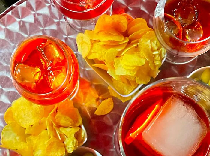 A gleaming metal table adorned with an array of cocktails in vibrant shades of red and orange, accompanied by bowls of potato chips and olives.