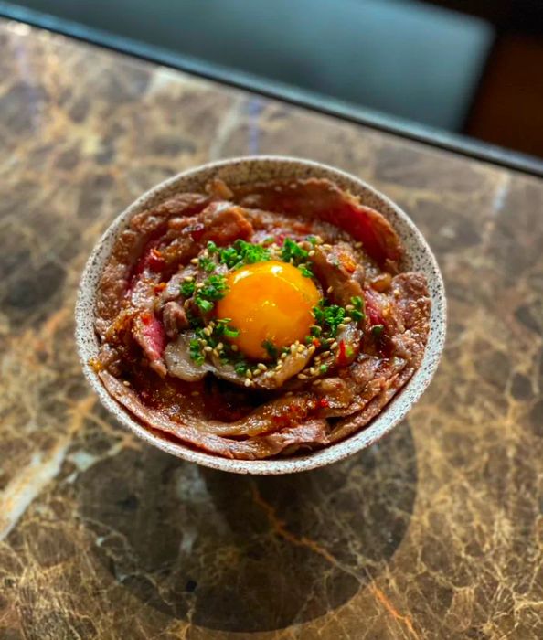 A petite bowl features curled strips of wagyu beef surrounding a vibrant egg yolk at the center, garnished with finely chopped herbs.