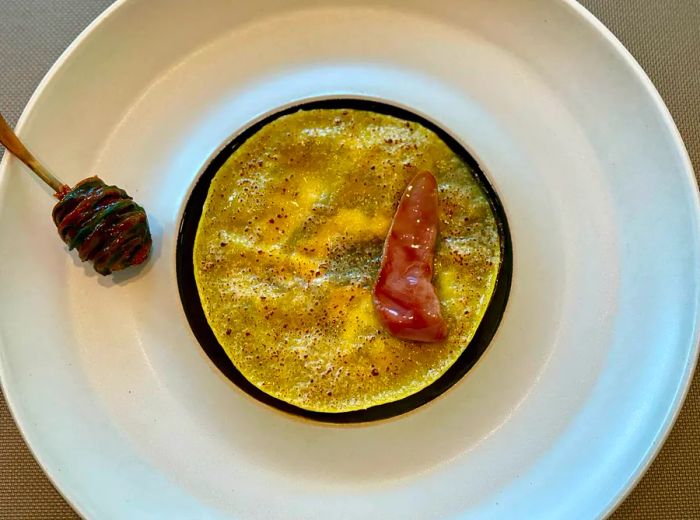 An aerial view of a circular lasagna topped with a strip of meat, elegantly presented on a large white plate accompanied by a honey stirrer.