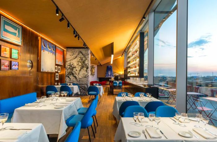 A restaurant interior featuring a glass wall offering a panoramic view of the city. The opposite wall showcases wood accents adorned with various artworks. Tables for four and six are elegantly set with white tablecloths, complemented by blue suede chairs.