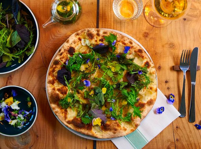 A pizza adorned with greens and flowers, accompanied by side dishes and wine.