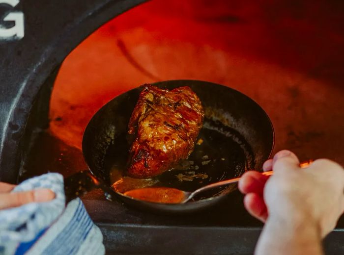 A chef bastes meat in a pan inside a wood-fired oven.