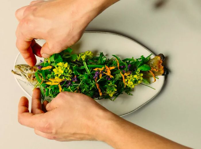 Hands delicately arrange a variety of sprigs and flowers atop a whole roasted fish.