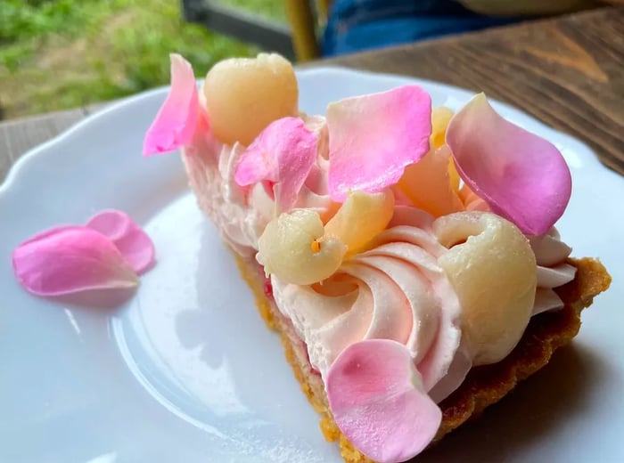 A slice of tart adorned with fruit, rose petals, and icing in soft white and pink hues.