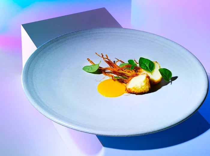 An elaborate dish featuring a halved root vegetable submerged in a vibrant yellow sauce, set against a geometric backdrop.