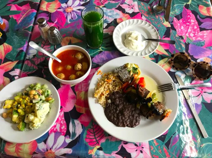 A vibrant table displays dishes including skewers, pilaf, “meatballs” in soup, and a fresh salad.