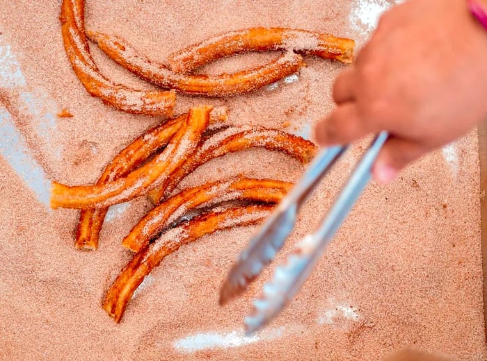 A tray filled with fried churros coated in cinnamon sugar, with a hand holding tongs poised above them.