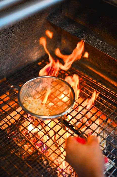 A hand holds a strainer filled with ant eggs above a flame.