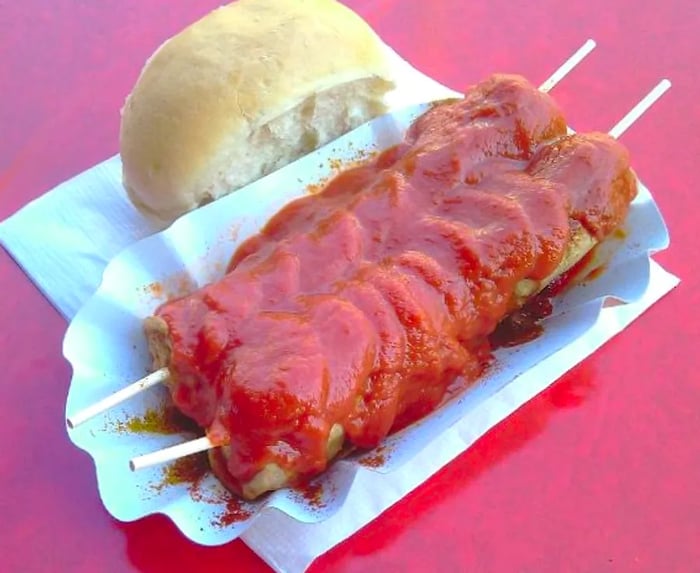 A pair of long sausages drenched in sauce, skewered and served in a paper boat alongside a roll on a red table.