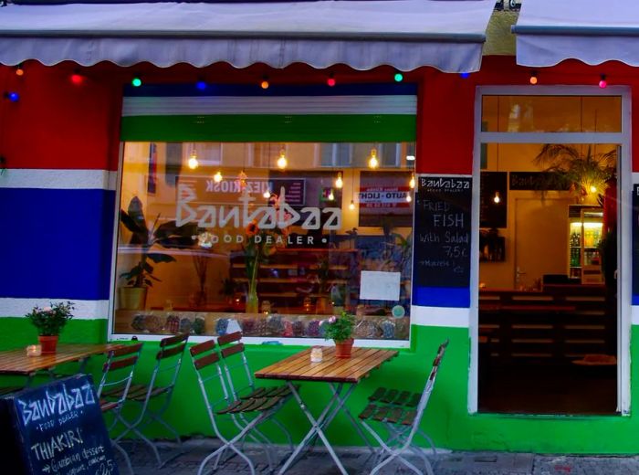 A vibrant restaurant facade featuring bold red, green, and blue paint under an awning, complemented by a few outdoor tables and chairs.