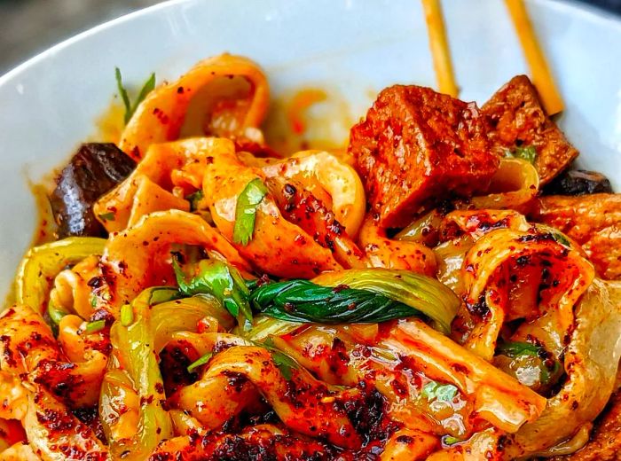 A bowl filled with thick noodles in a spicy chili sauce, topped with tofu and stir-fried vegetables.