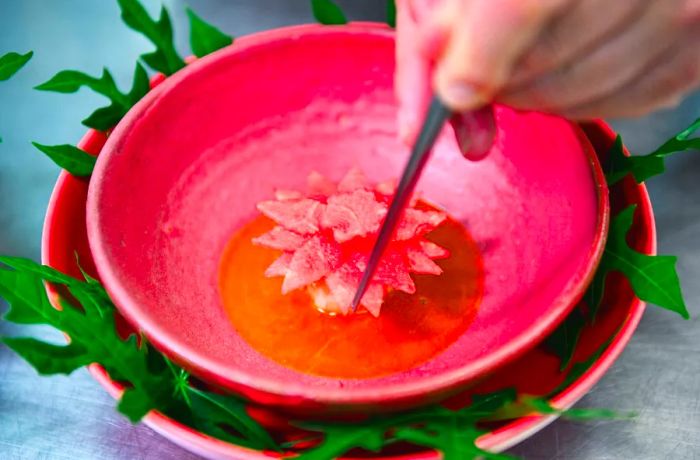 An unseen chef delicately finishes a vibrant pink dish that resembles a blooming flower using tweezers.