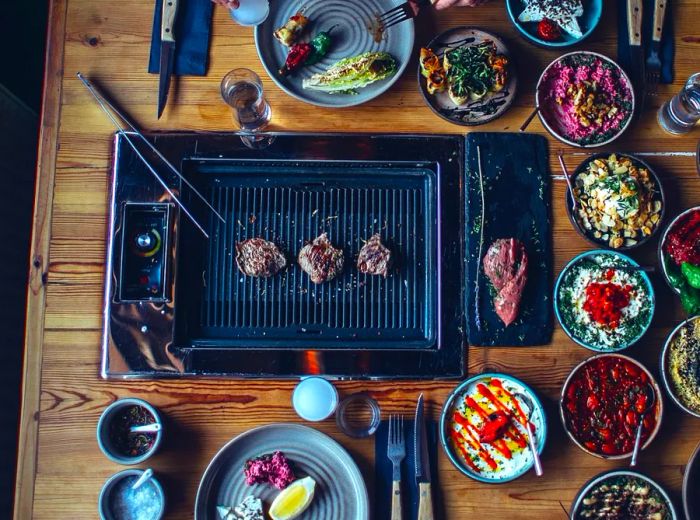 An overhead view of a table featuring a grill at its center, where meat sizzles. Surrounding the grill are numerous small dishes filled with vibrant ingredients. A diner wearing a black hat occupies one end of the table.