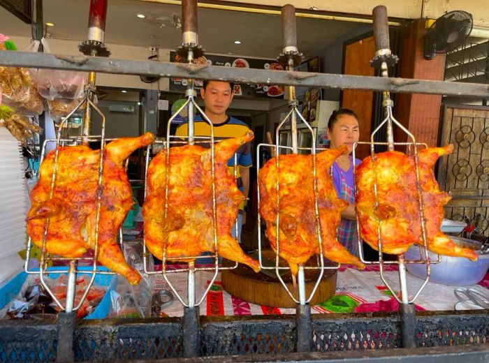 Workers are positioned behind a row of chickens skewered and grilling on spits.