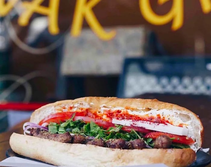 An elongated sandwich opened up to display meat, chopped herbs, tomato, onion, and sauce.