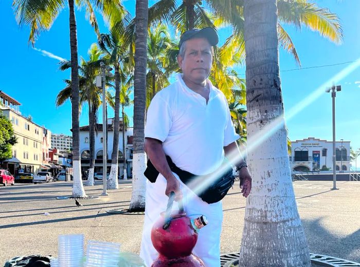 A vendor stands under the sun with a jug and drink supplies on a concrete path flanked by palm trees.