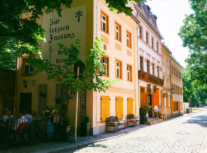 A charming corner building on a sunny cobblestone street, adorned with bright yellow shutters and surrounded by lush foliage.