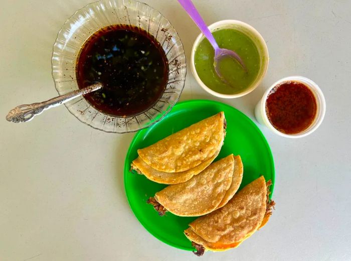 Three tacos artfully arranged with an assortment of sauces and a cup of consomé.