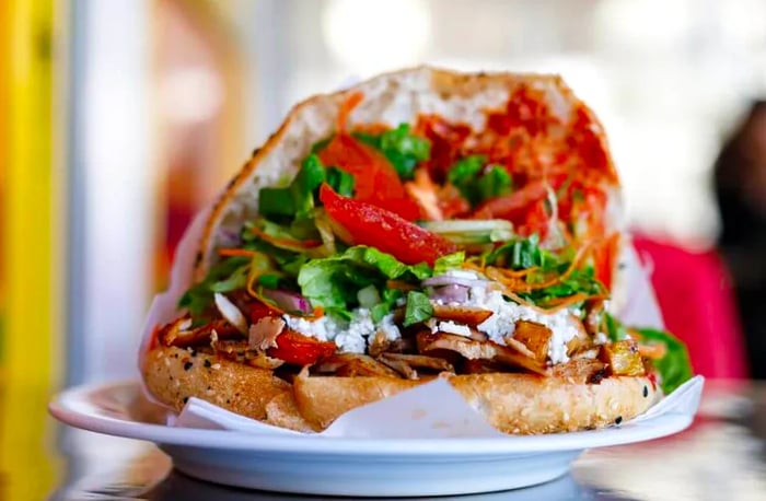 A flatbread sandwich filled with slices of grilled meat, heaped with greens, tomatoes, herbs, onions, and sauce.