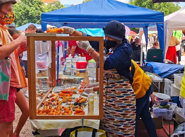 A vendor passes an item over the glass display of a makeshift food stall in a sunny park to a customer on the other side.