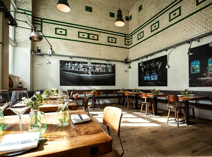 A spacious, sunlit dining area featuring large photographs on the walls, wooden tables set for lunch, pendant lighting, and subway-tiled walls.