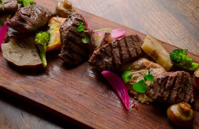 From above, a beautifully arranged meat platter featuring various cuts of grilled steak adorned with char marks, alongside hearty portions of cooked vegetables and fresh herbs for garnish.