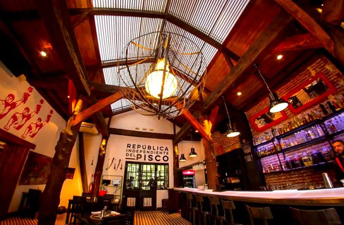 A low-angle view captures an expansive bar illuminated by a large skylight above. The scene features a central caged pendant light, tiled flooring, several tables, tall shelves backlit with light, and a bartender busy at work.