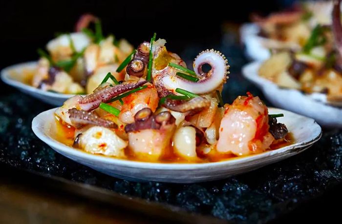 A bowl brimming with chunks of assorted seafood in a flavorful broth, with similar dishes softly blurred in the background on a dark countertop.