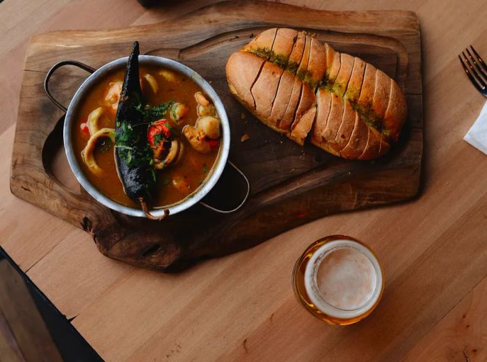 From an overhead view, a wooden table showcases a meat board alongside a bowl of seafood and vegetable stew, topped with a large pepper resting on the rim, accompanied by a loaf of bread smeared with sauce and sliced.