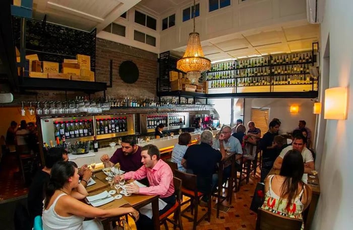 Guests relax at tables in a warm and inviting bar, adorned with pendant lights, lined with rows of bottles along the bar and up the back wall, and featuring dark tiled floors.