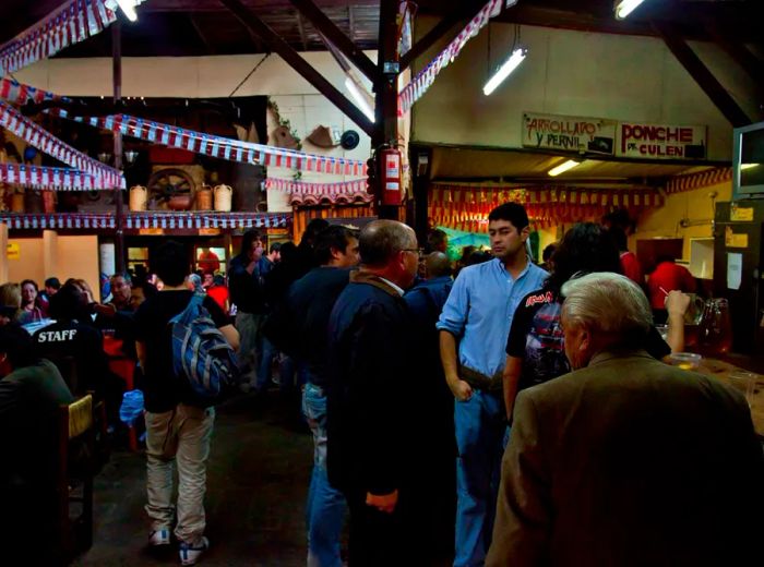 A lively crowd gathers at a bar in a spacious venue featuring slender wooden support beams draped with colorful streamers, surrounded by tables full of patrons.