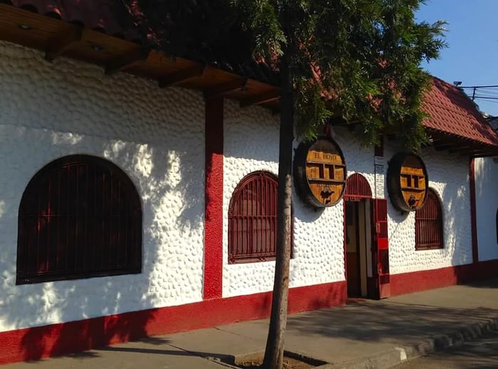 A single-story building featuring a shingled roof, textured white walls, large arched windows secured with heavy bars, and barrels flanking the entrance.