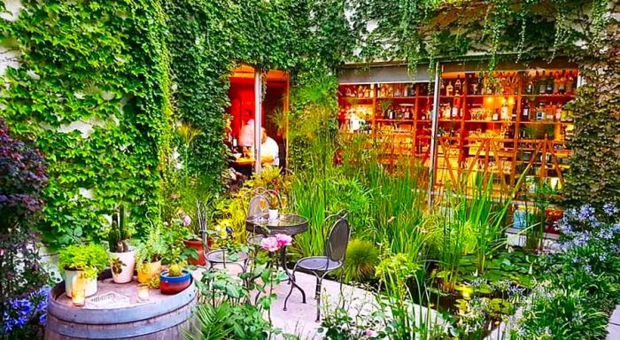 A vibrant outdoor patio lush with greenery, featuring a striking plant wall and cascading vines overhead, with cozy patio chairs and a library-like interior visible through expansive windows.