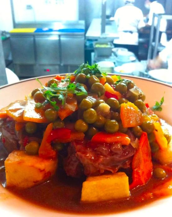 A close-up view of a plate featuring steak adorned with peas, carrots, and an assortment of roasted vegetables, all resting in a rich, thick sauce that collects in the dish.