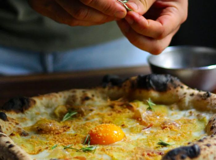 A chef sprinkles fresh herbs onto a pizza, showcasing a vibrant yellow egg yolk at its center.