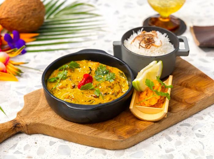 A cast iron dish filled with vibrant yellow coconut curry, garnished with herbs and a red pepper, served alongside a pot of rice and various accompaniments.