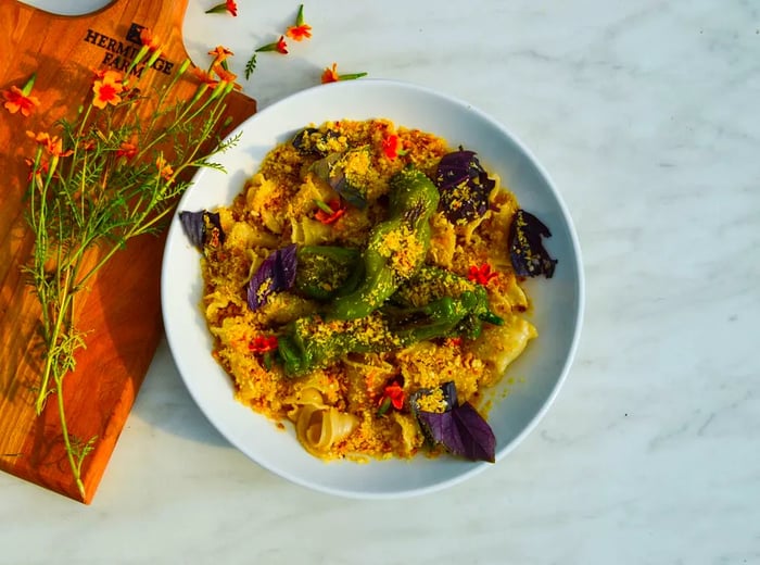 A bird's-eye view of a pasta dish garnished with large, charred shishito peppers and breadcrumbs, placed on a marble countertop adorned with flowers.