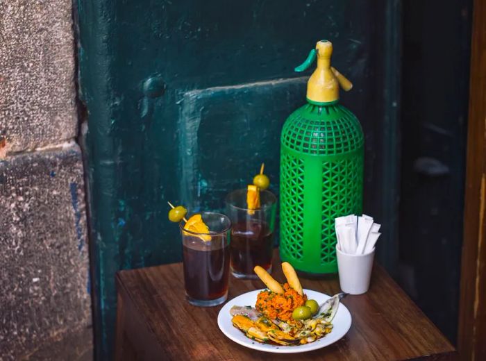 A rustic wooden table holds a plate of vibrant tapas, accompanied by two glasses of vermouth garnished with olives and orange slices on skewers.