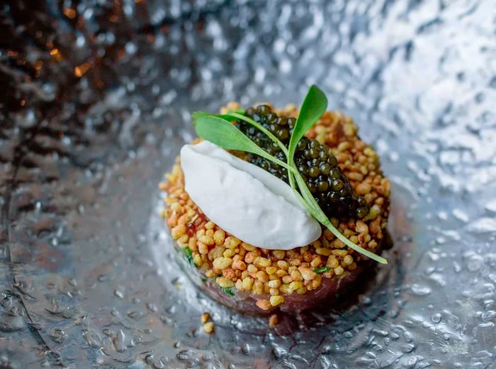 A glossy black dish featuring a small round tuna patty adorned with buckwheat, a dollop of cream, a heap of caviar, and a sprig of herbs for decoration.