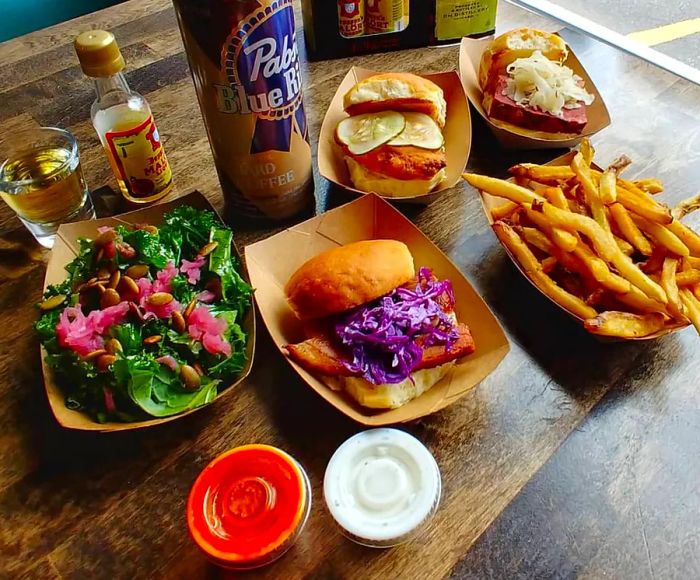 Paper boats filled with dishes sit on a wooden table next to a can of Pabst, featuring an assortment of sliders—one topped with bright purple cabbage—accompanied by a salad and fries.