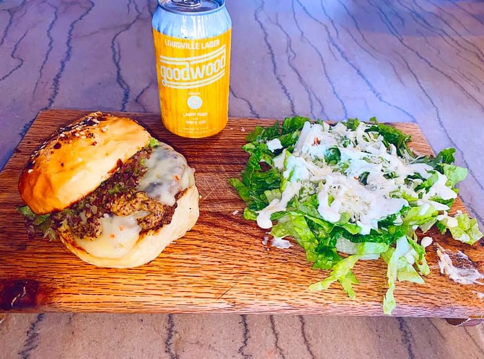 A burger topped with melted cheese, the top bun slightly lifted, served on a wooden plank alongside a dressed salad and a nearby can of beer.