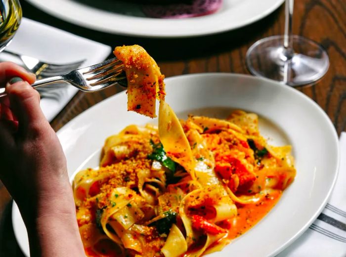 A diner showcases a thick noodle from a plate of pasta drenched in red sauce and loaded with vegetables.