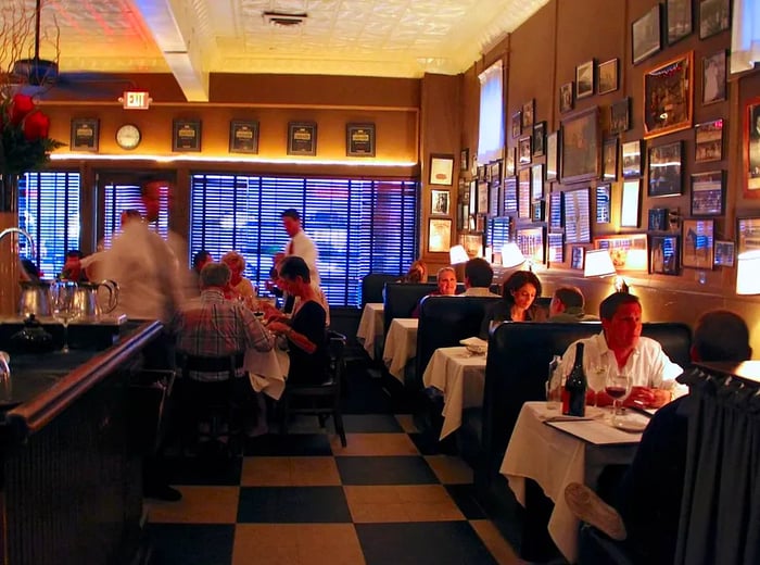 An inviting restaurant interior features diners at small tables, walls adorned with framed photographs, servers bustling about, a checkered floor, and windows with drawn shades.