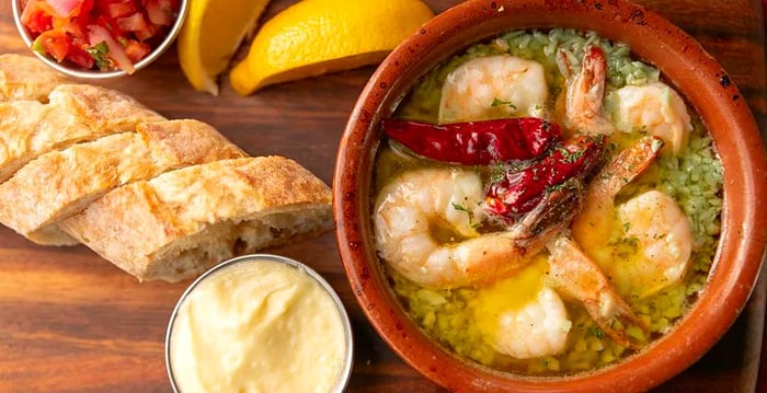 A top-down view of a ceramic bowl brimming with shrimp in garlic sauce, accompanied by slices of bread, dipping sauce, a cup of salsa, and wedges of lemon.